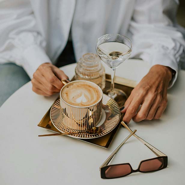 Person at cafe table with elegant coffee cup, glass of water, and sunglasses.