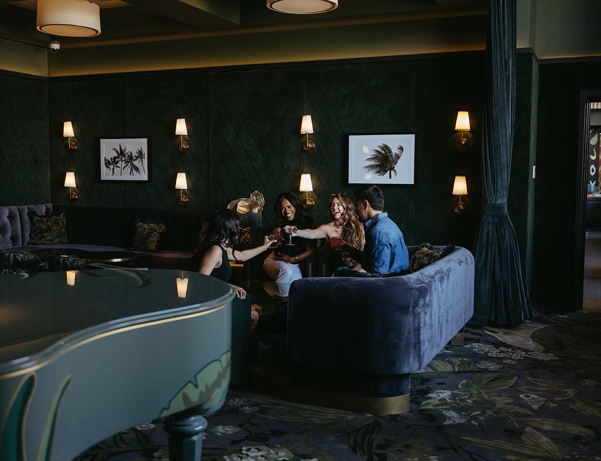 Friends toasting with cocktails in dark, elegant bar with plush seating and piano.
